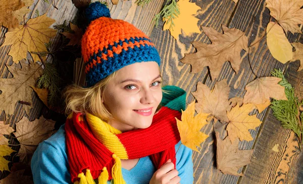 Knitted accessories. Fall cozy atmosphere. Girl cheerful face lay on wooden background with leaves top view. Fall and autumn season concept. Hipster woman knitted hat and scarf hold autumn leaves