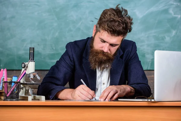 Учитель бородатый человек пишет с ручкой, заполненной бумажной работой. Учитель формальная куртка сидеть стол классной доски фона. Письменный отчет. Ежедневная рутинная работа учителей и написание отчетов — стоковое фото