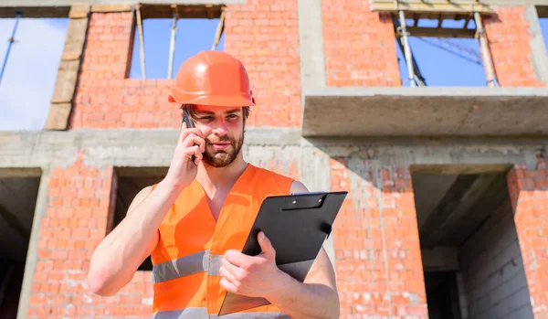 Engineer phone call control process. Guy protective helmet stand in front of building made out of red bricks. Builder vest and helmet works at construction site. Contractor control according to plan