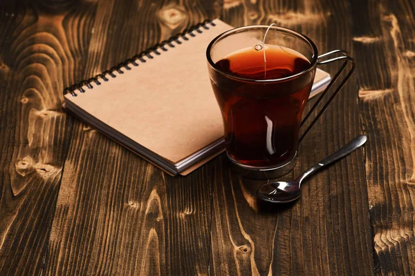 Tee und Notizbuch auf Holzgrund. Zusammensetzung von Tasse und Lehrbuch zum Entspannen. Herbstliches Abendkonzept. Glas schwarzer Tee mit Teebeutel neben Teelöffel und geschlossenem Buch — Stockfoto