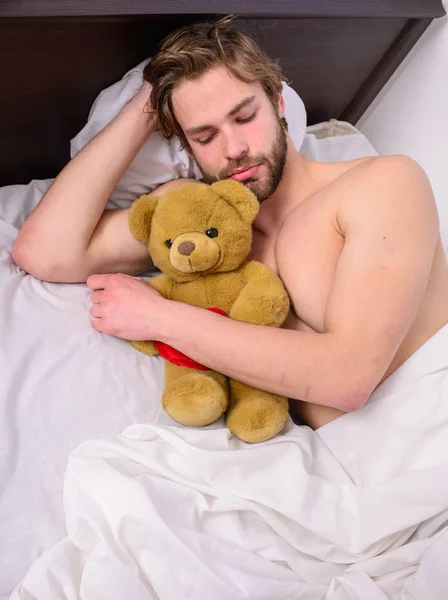 Sweet dreams concept. Sweet memories from childhood. Guy lay white bedclothes with toy. Man unshaven bearded face relax with favorite teddy bear. Man handsome guy relaxing bed hug teddy bear toy
