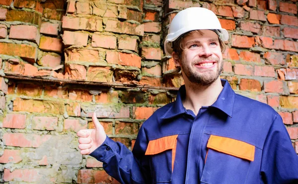 Guy beschermende helm bebaarde knappe bouwer. Bouwer helm bouw site bakstenen muur achtergrond. Man neemt pauze werkdag op de bouwplaats. Guy glimlachend toont duimschroef opwaarts gebaar — Stockfoto