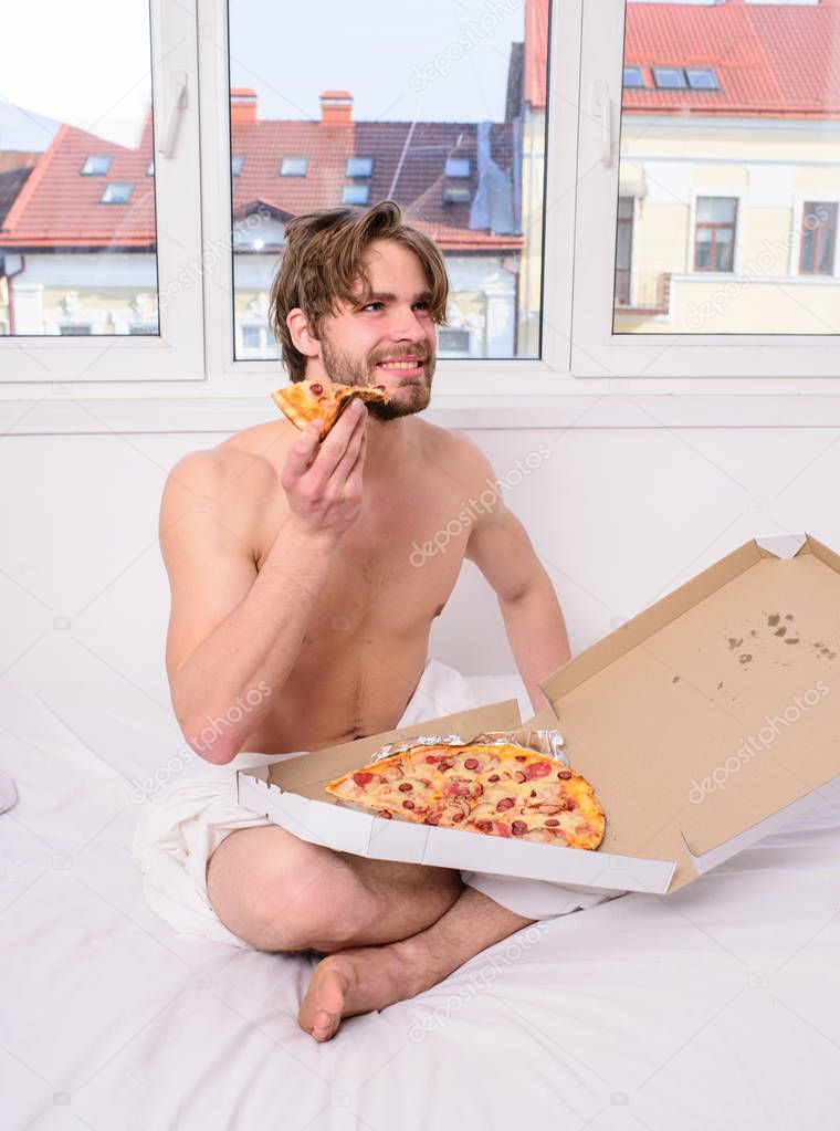 Break diet concept. Food delivery service. Man bearded handsome bachelor eating cheesy food for breakfast in bed. Man likes pizza for breakfast. Guy holds pizza box sit bed in bedroom or hotel room