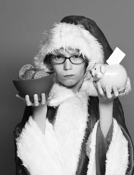 Joven lindo santa claus chico con gafas en suéter de Navidad y año nuevo sombrero de Navidad celebración de la hucha de cerdo rosa y galletas de chocolate de la cadera en tazón sobre fondo rojo estudio —  Fotos de Stock