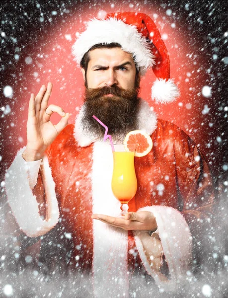 Bonito barbudo santa claus homem com longa barba no rosto sério segurando vidro de coquetel não alcoólico no Natal ou xmas suéter e chapéu de ano novo mostrando ok no fundo estúdio vermelho — Fotografia de Stock