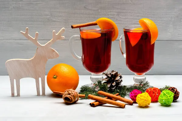 Mulled wine or hot beverage with cinnamon sticks, orange fruit and fir cone. Glasses with mulled wine or hot drink near wooden deer decoration on white background, close up. Winter beverage concept — Stock Photo, Image