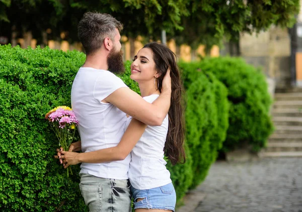 Verliefde paar hugs buiten park achtergrond. Man bebaarde hipster hugs zachtjes vriendin genieten van haar zachte lange haren. Hij houdt van haar prachtige haar. Meisje houdt bloemen terwijl man strelen haar lange haren — Stockfoto