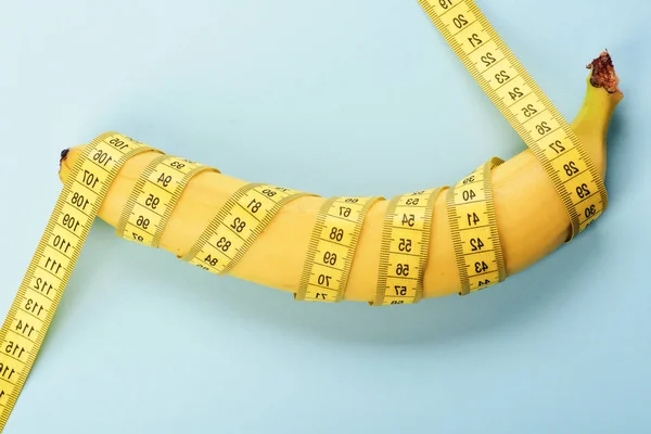 Regime, healthy food and weight management concept. Centimeter ruler spinned around fresh fruit. Yellow tape around banana isolated on light blue background. Banana with tape for measuring figure.