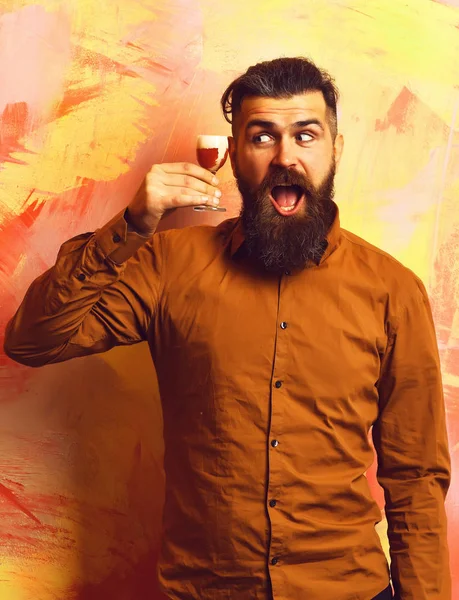 Bearded man, long beard. Brutal caucasian funny happy hipster with moustache in brown shirt holding alcoholic red shot on colorful texture background.
