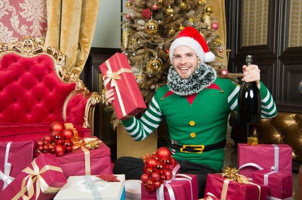 Man dragen santa hat elf kleren vieren Kerstmis interieur versierd fir tree en geschenken. Kerst champagne drinken. Man vieren Nieuwjaar of kerstvakantie. Santa en kerst cadeaus concept — Stockfoto