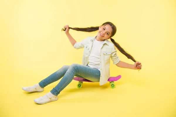 Girl show long ponytail hairstyle sit penny board yellow background. Child cute hairstyle ride penny board. Proud of long hair. Hairstyle for active leisure. Keep hairstyle tidy during sport activity