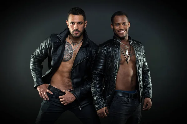 Men on smiling faces with bristle. Leather masculine clothing concept. Machos with muscular torsos look attractive in leather jackets, dark background. Men with sexy muscular torsos look brutally
