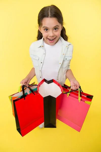 Check out her profitable purchases. Girl carries shopping bags yellow background. Girl fond of shopping. Child cute shopaholic with bunch shopping bags black friday total sale. Exciting shopping — Stock Photo, Image