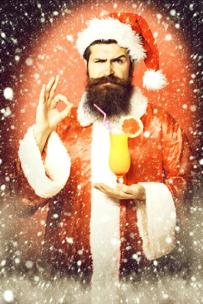 Stilig skäggiga santa claus man med långt skägg på allvarliga ansikte holding glas alkoholfria cocktail i jul eller xmas tröja och nyår hatt visar ok på röda studio bakgrund — Stockfoto