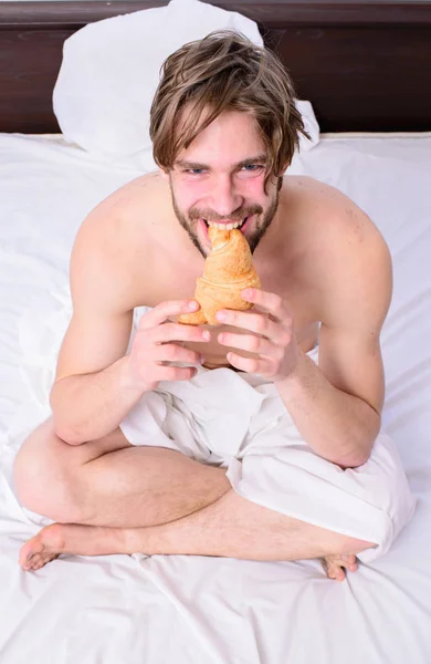 French breakfast stereotype. Man eats croissant he likes bakery products. Fresh bakery product. Man bearded handsome guy eating breakfast in bed. Guy holds croissant sit bed in bedroom or hotel room