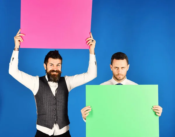 Advertisement or commercial posters. Announcement or promotion concept. Men with beards hold boards on blue background. Businessmen with strict and happy faces hold green and pink boards, copy space