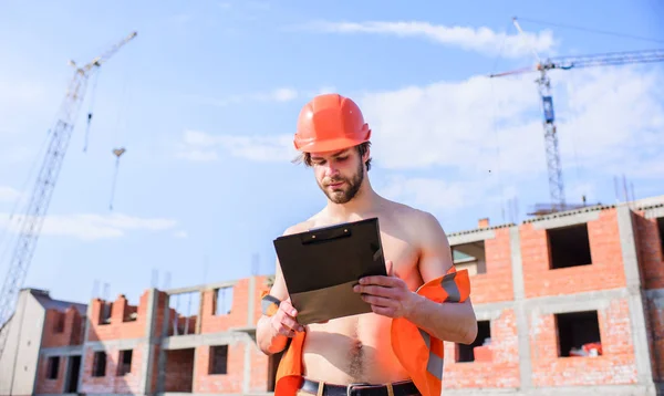 Kontroll byggprocessen. Entreprenören kontroll enligt plan. Guy skyddande hjälm stå framme av byggnad gjord av rött tegel. Builder orange väst och hjälm fungerar på byggarbetsplats — Stockfoto