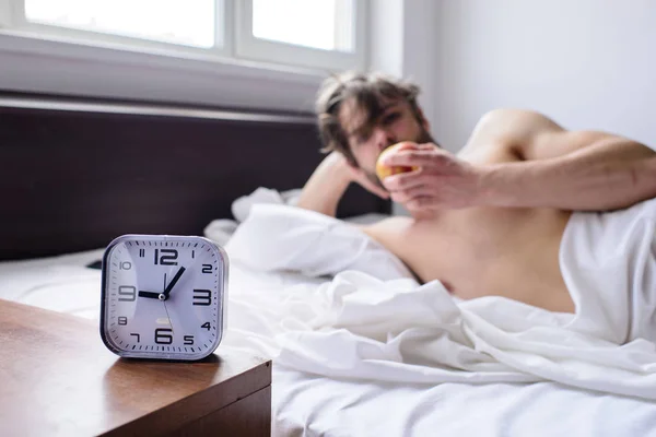 Get up early morning tips. Man bearded unshaven handsome guy lean on hand while eats fruit near alarm clock defocused. Guy relaxing bed before alarm clock ringing. Important part of morning