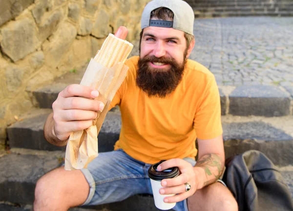 Élvezze az étkezés koncepció. Városi élelmiszer-kultúra fogalma. Csípő enni hot-dog ül míg kint a lépcsőn. Szakállas embert csípő élvezze utcai élelmiszer-városi háttér. Finom hot dog kolbász a roasty zsemle — Stock Fotó