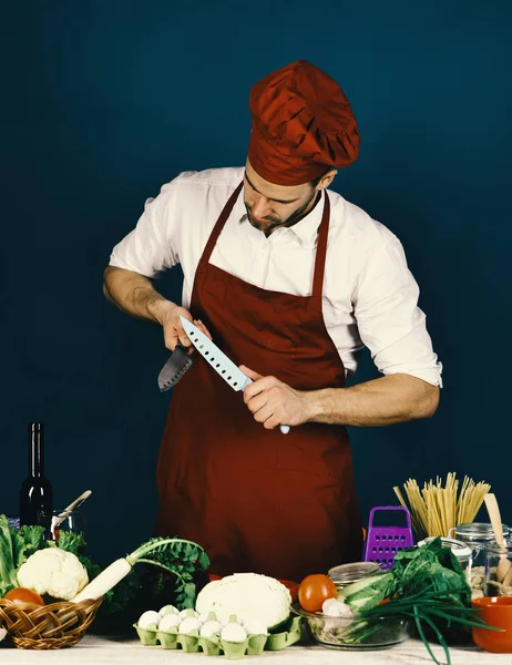 Hombre en sombrero de cocina y delantal afilan cuchillas . — Foto de Stock