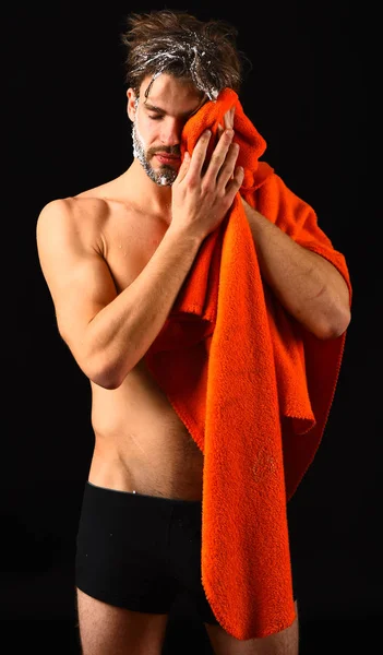 Man with orange towel wipe face, keep soap suds out of eyes. Macho attractive nude guy black background. Man bearded tousled hair covered with foam or soap suds. Wash off foam with water carefully