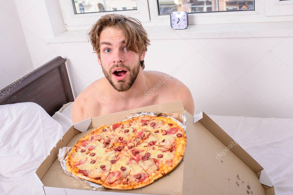 Man bearded handsome guy eating cheesy food for breakfast in bed. Guy holds pizza box sit bed in bedroom or hotel room. Food delivery service. Man likes pizza for breakfast. Who cares about diet
