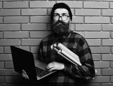 Sakallı adam, uzun sakallı. Kafkas asıllı, traşsız bir hippi dizüstü bilgisayar ve kağıt rulo kağıtlar taşıyor. Kırmızı siyah kareli gömlek, şapka ve gözlüklerle kahverengi tuğla duvarlı stüdyo arka planında.