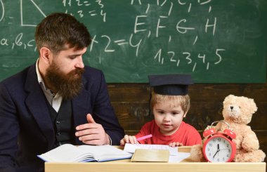 İlköğretim. Öğretmenle okuyan bir çocuk. Baba oğula öğretir, tartışır, açıklar. Eğitim kavramı. Sınıfta harç panosunda resmi giyim öğretmeni ve öğrenci, arka planda tebeşir tahtası