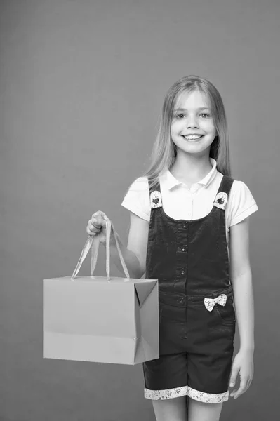 Маленький ребенок с пакетом покупок на фиолетовом фоне. Счастливая девушка улыбается бумажным пакетом. Покупатель в комбинезоне. Праздники и праздники. Удовлетворена покупками. продажа и черная пятница — стоковое фото