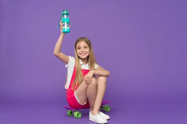 Оставаться обезвоженным. Девушка счастливое лицо держит с бутылкой воды в то время как сидеть пенни доска фиолетовый фон. Девочка заботится о гидратации тела. Активный отдых и водный баланс. Активный и здоровый ребенок пьет воду — стоковое фото