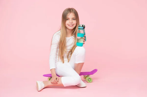 女の子の幸せそうな顔を座っているペニー ボード ピンク背景中の水のボトルを保持します。子供女の子ケア体水分補給します。アクティブなレジャーと水のバランス。アクティブで健康的な子供は、水を飲む。水和滞在 — ストック写真