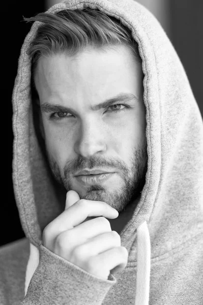 Guy bearded and attractive touches bristle on chin. Masculinity concept. Man with beard or unshaven guy looks handsome hooded. Man with bristle on serious face, urban background, defocused