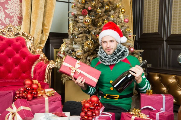 Счастливый человек с подарочной коробкой и шампанским на рождественской елке. Мужчина в костюме Санта Клауса с рождественским подарком. Зажги елку. Это рождественский день, давайте распространять хорошее настроение — стоковое фото