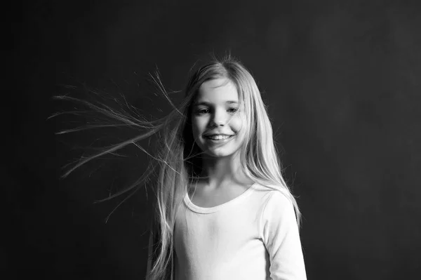 Moda, mira peinado. Modelo de niño sonriendo con el pelo largo que sopla. Chica con sonrisa adorable sobre fondo oscuro. Concepto de infancia feliz. Salón de belleza y peluquería . — Foto de Stock