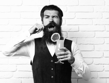 Uzun sakallı ve bıyıklı yakışıklı adamın komik yüzünde şık saçları var. Elinde beyaz tuğla duvar arka planında klasik süet deri yelek ve alkollü içecek bardağı var.