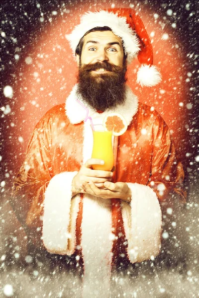 Guapo barbudo hombre de Santa Claus con barba larga en la cara sonriente sosteniendo vaso de cóctel sin alcohol en navidad o suéter de Navidad y sombrero de año nuevo en fondo rojo estudio —  Fotos de Stock