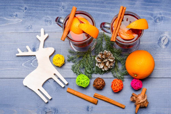 Mulled wine or hot beverage with cinnamon, orange fruit and fir cone, top view. Winter beverage concept. Glasses with mulled wine or hot drink near wooden deer decoration on blue wooden background — Stock Photo, Image