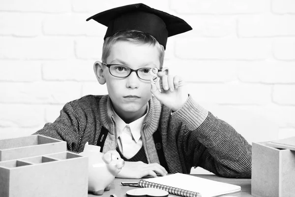 Ung söt elev pojke i grå tröja och glasögon sitter vid skrivbordet med copybook trä siffror rosa gris bank i examen mössa i klassrummet på vit tegelvägg bakgrund — Stockfoto
