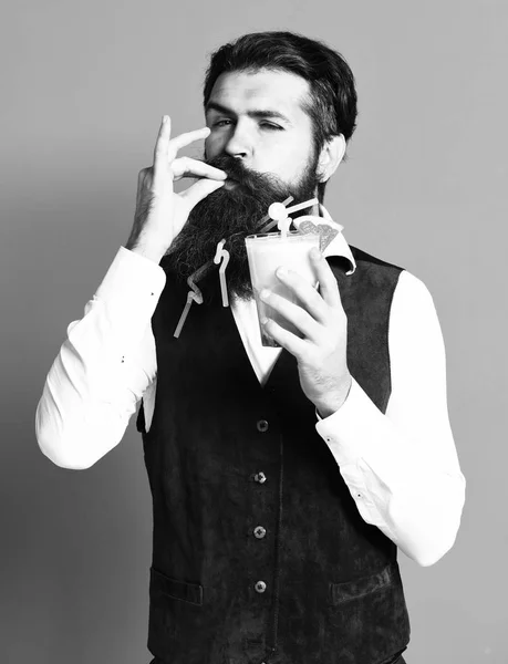 Bel homme barbu avec longue barbe et moustache a des cheveux élégants sur le visage drôle tenant verre de boisson alcoolisée en daim vintage gilet en cuir sur fond de studio rouge — Photo