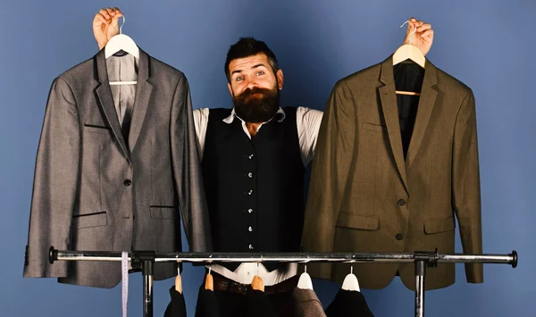 Designer makes choice near clothes hangers. Tailor with confused face holds grey suits near custom jackets on blue background. Fashion and individual style concept. Man with beard by clothes rack.