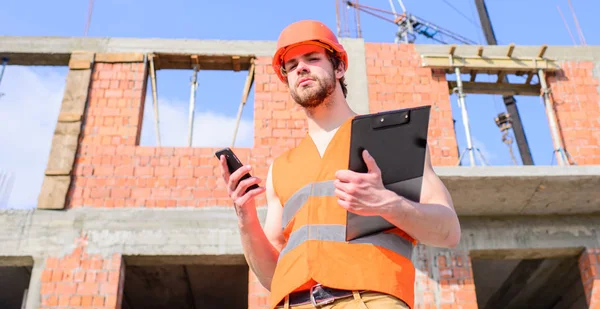 Ingenjören telefonsamtal kontroll process. Entreprenören kontroll enligt plan. Guy skyddande hjälm stå framme av byggnad gjord av rött tegel. Builder väst och hjälm fungerar på byggarbetsplats — Stockfoto