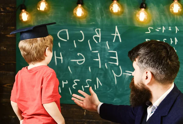 Boy, child in graduate cap looks at scribbles on chalkboard while teacher explains. Teacher with beard, father teaches little son in classroom, chalkboard on background. Basics of education concept — Stock Photo, Image