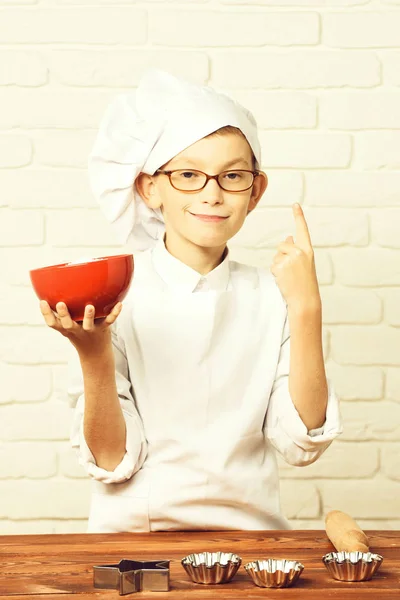 Niño pequeño cocinero lindo en uniforme blanco y sombrero en la cara divertida con gafas de pie cerca de la mesa con moldes de rodillo para pasteles y la celebración de tazón rojo en el fondo de la pared de ladrillo — Foto de Stock