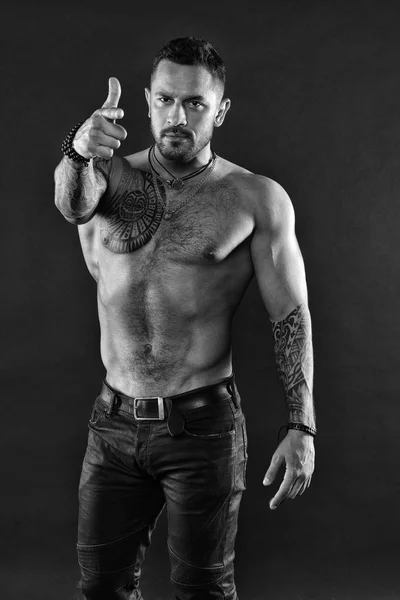 Tattoo art. Handsome fit tattoed man wear jeans pointing forward. Muscular tattooed athlete look attractive. Sport and fashion concept. Man handsome shirtless muscular with jeans over dark background