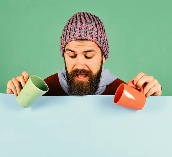 Breakfast time idea. Perfect autumn morning and aroma concept. Man in warm hat holds orange and green cups on green and cyan background, copy space. Hipster with beard and curious face looks down