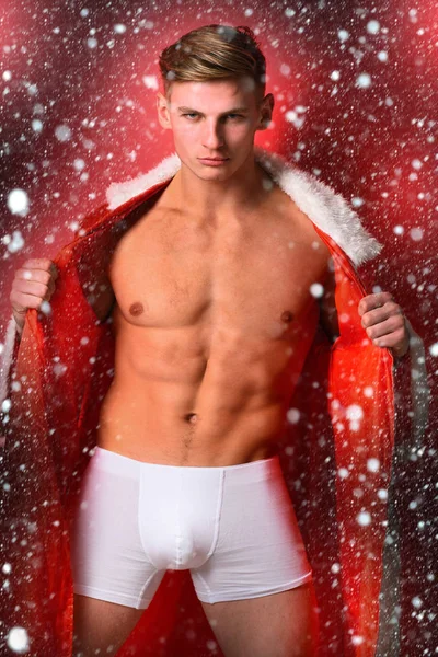 Joven guapo macho santa man con sexy muscular atlético fuerte cuerpo ha desnudo torso y fuerte vientre en navidad o Navidad suéter en rojo estudio fondo — Foto de Stock