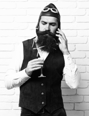 Sakallı pilot yakışıklı uzun sakal ve bıyık ciddi yüz alkolik bardak tutan vintage süet deri yelek ateş ve beyaz tuğla duvar arka plan üzerinde cep telefonuyla konuşuyor