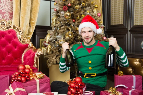 Man vieren Nieuwjaar of kerstvakantie. Sinterklaas en kerst geschenken concept. Man dragen santa hat elf kleren vieren Kerstmis interieur versierd fir tree en geschenken. Magische kerstperiode — Stockfoto