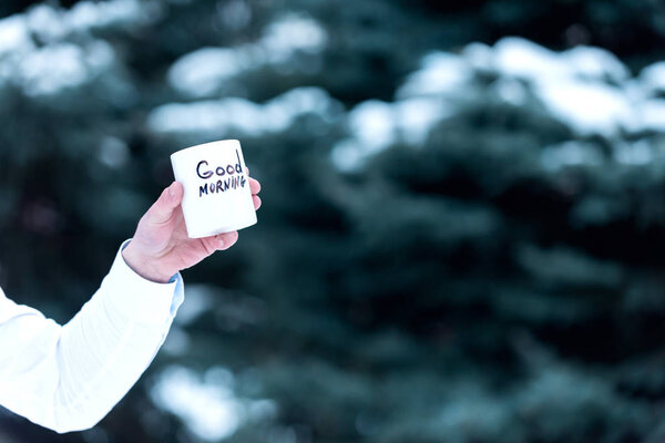 Hot beverage on nature background, defocused. Cup of tea or coffee with Good Morning inscription. Male hand holds white mug with drink. Morning drink outside concep