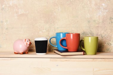 colorful cups on book and pink piggy banks and coffee cup on beige textured and wooden background, copy spac clipart